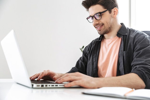 Side view of Smiling handsome man in eyeglasses using laptop computer while sitting by the table at office