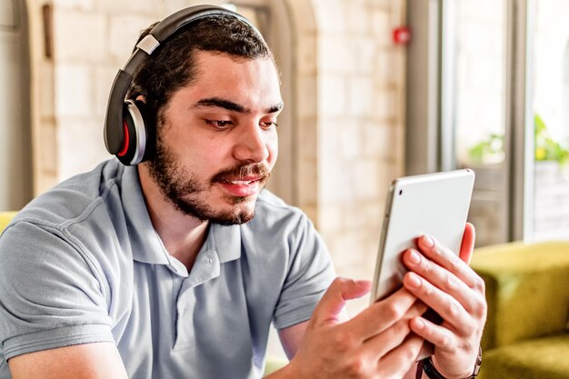 Side view of smiling man with headphones using a tablet sitting on a couch at home