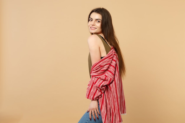 Side view of smiling pretty young woman in casual clothes standing and looking camera isolated on pastel beige wall background in studio. People sincere emotions lifestyle concept. Mock up copy space.