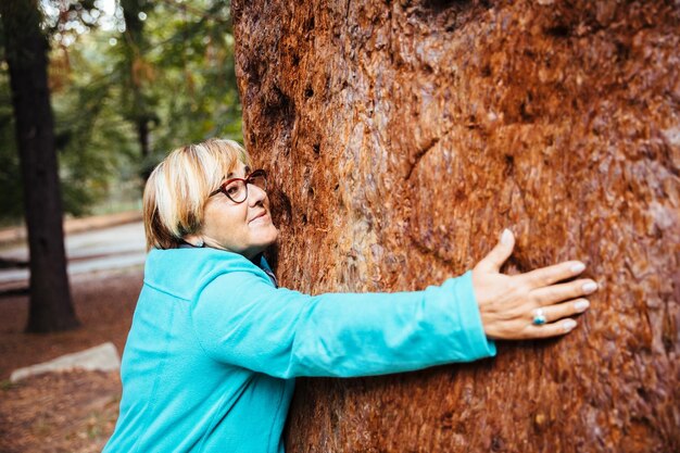 Side view of smiling senior woman hugging tree trunk