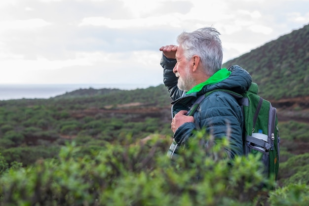 Side view of a smiling white-haired elderly man in healthy excursion among mountain and the sea. Horizon over the water. Active retiree backpacker enjoying the outdoors and freedom