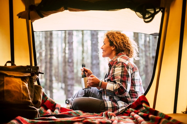 Боковой вид улыбающейся женщины с кружкой в руках, сидящей у палатки в лесу