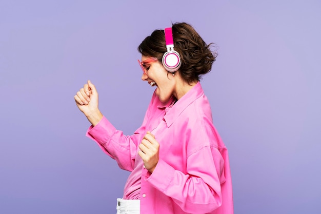 Боковой вид улыбающейся женщины в розовой рубашке, слушающей музыку в наушниках, танцующей