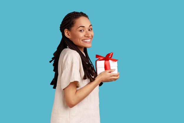 Вид сбоку улыбающейся женщины с дредами, держащей подарочную коробку, поздравляющую с подарком, смотрящую в камеру в белой рубашке. Снимок в студии на синем фоне