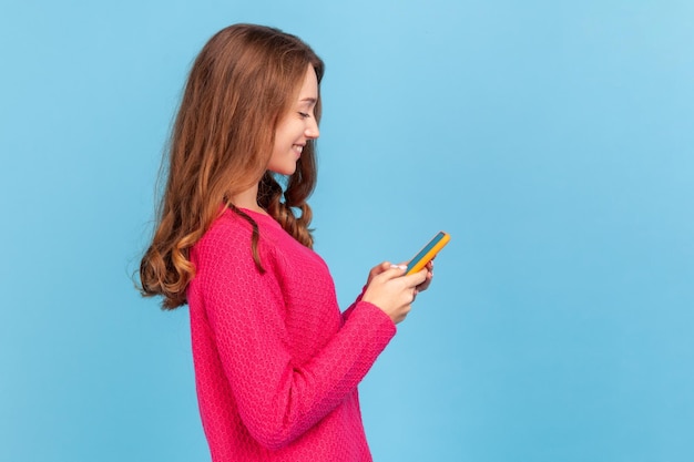손에 스마트 폰을 들고 서 있는 분홍색 풀오버에 물결 모양의 머리를 한 웃는 여성의 측면 보기, 스크롤링, 인터넷 검색, 소셜 네트워크 확인. 파란색 배경에 고립 된 실내 스튜디오 촬영입니다.