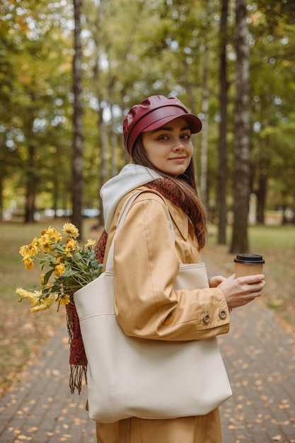 Photo side view of smiling young female with cup of coffee to go standing in park in fall season