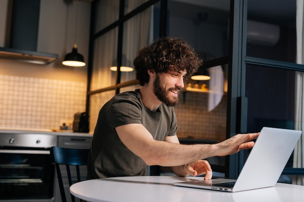 Side view of smiling young freelancer male opening laptop and starts typing on keyboard sitting at table in kitchen with modern interior