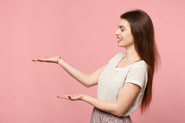 Side view of smiling young woman in casual light clothes posing isolated on pink wall background, studio portrait. People sincere emotions lifestyle concept. Mock up copy space. Pointing hands aside.