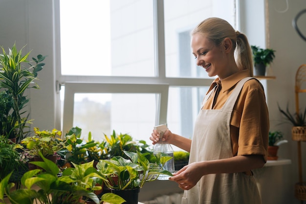 噴霧器によって植木鉢の観葉植物に水を噴霧するエプロンで笑顔の若い女性の花屋の側面図
