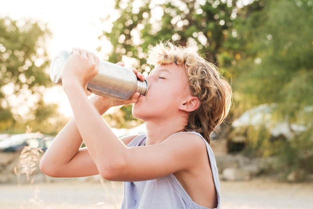 Side view of sporty boy in sportswear drinking fresh water from bottle while resting in outdoor park