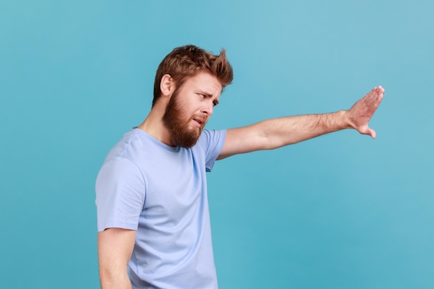 Side view of strict bearded man showing stop sign standing with frowning face and bossy expression meaning caution to avoid danger or mistake Indoor studio shot isolated on blue background