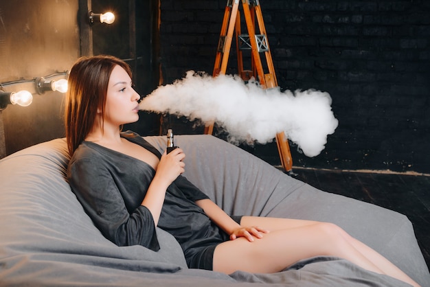 Side view of stunning young woman using an electronic cigarette, exhaling big cloud of smoke, spending leisure time while relaxing on the poof in the room with stylish interior.