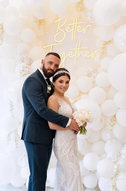 Side view of stylish bridegroom with beard and mustaches in trendy suit standing and hugging bride which smiling and looking at camera while posing with him on wedding arch background