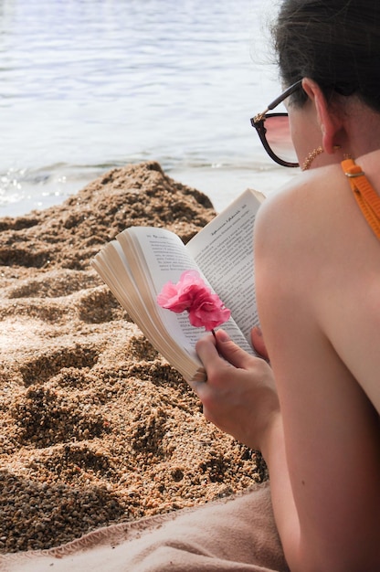 La vista laterale della donna alla moda in occhiali da sole sta leggendo un libro sulla spiaggia di sabbia