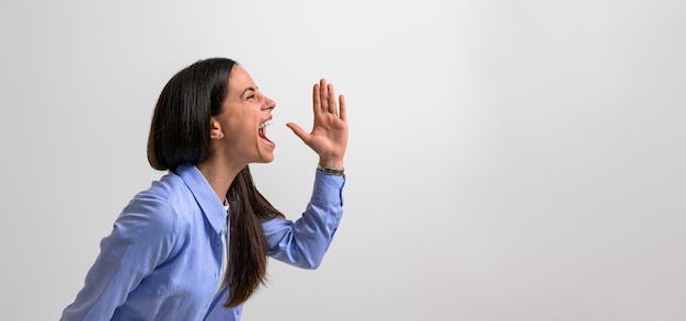 Side view of successful female professional screaming and gesturing in joy while standing isolated on background Young businesswoman dressed in casuals showing hand sign and shouting
