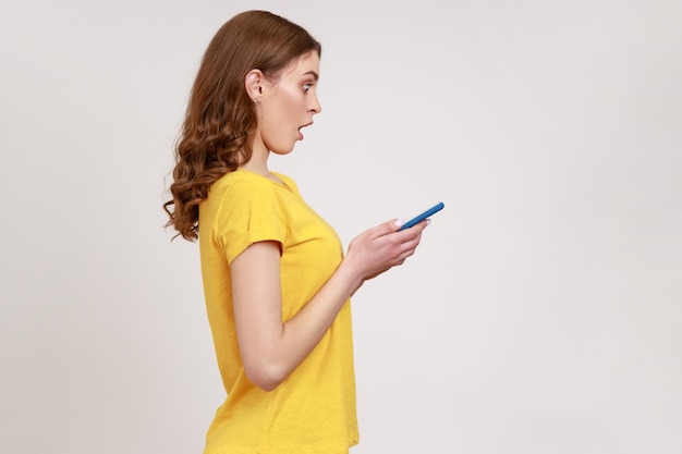 Side view of surprised teenager girl in yellow casual T-shirt, expressing shock while using cell phone, searching web, astonishing news. Indoor studio shot isolated on gray background.