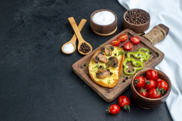 Вид сбоку вкусной закуски с грибами и нарезанными овощами на деревянной доске, набор столовых приборов, специй на черном фоне