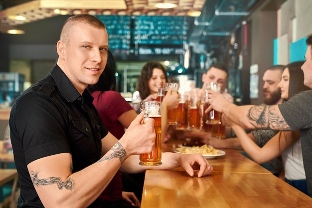 Side view of tattooed man in black shirt keeping glass of beer, looking at camera and posing in pub. Young strong man smiling and drinking tasty ale with friends in bar. Concept of hun and leisure.