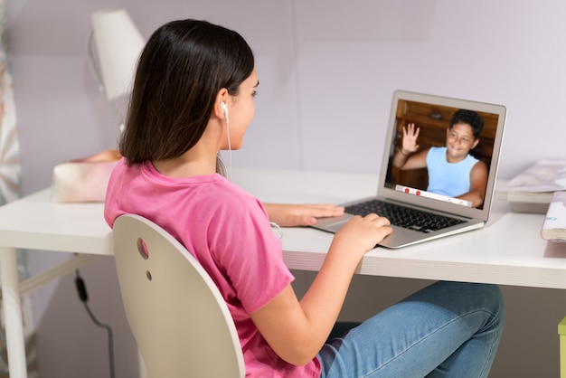 Side view of teen girl in earphones sitting at table and communicating via video chat app on laptop during social distancing period at home