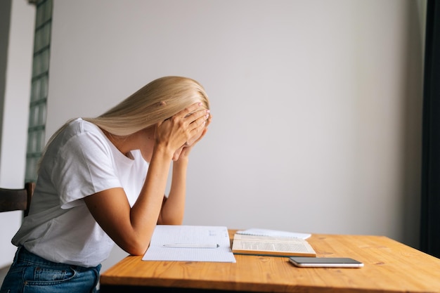 Side view of tired blonde young woman massaging temples suffering from headache after paperwork sitting at desk with book and notebook at home