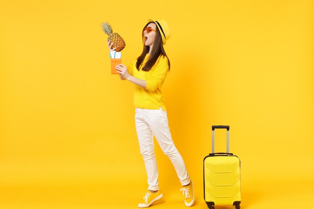 Side view of traveler tourist woman in hat hold passport tickets fresh pineapple fruit isolated on yellow orange background. Passenger traveling abroad on weekends getaway. Air flight journey concept.