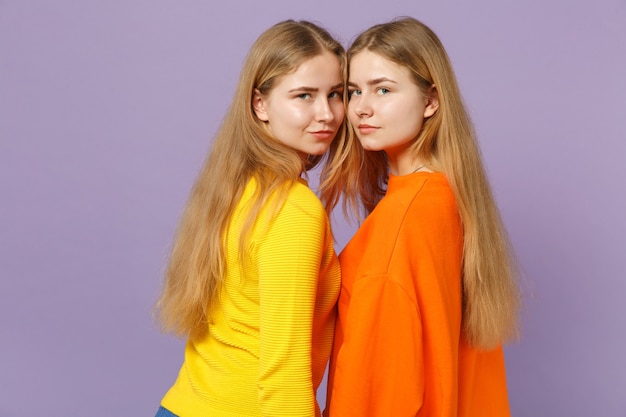 Side view of two beautiful young blonde twins sisters girls in vivid colorful clothes looking camera isolated on pastel violet blue wall. People family lifestyle concept. 