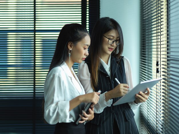 Side view of two businesswomen consulting on their work while standing next to window in office