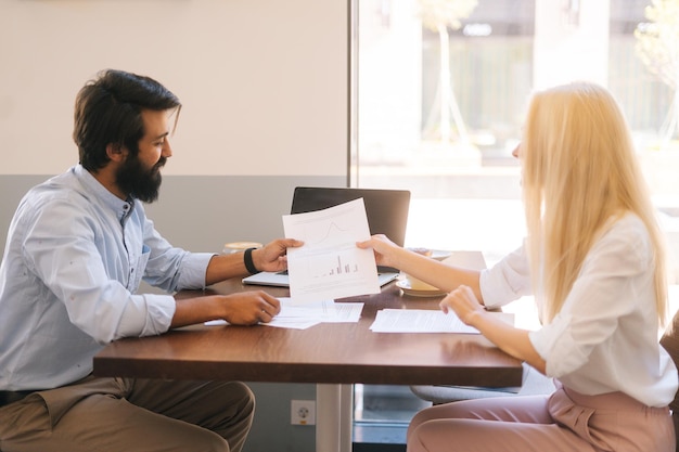 Side view of two multiethnic colleagues discuss financial data in report holding documents and charts sitting at desk in cafe by window Male and female business partners having meeting in coffee shop