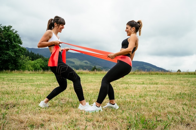 Side view of two women doing strength exercises with a rubber band, doing a lot of effort in the middle of the forest