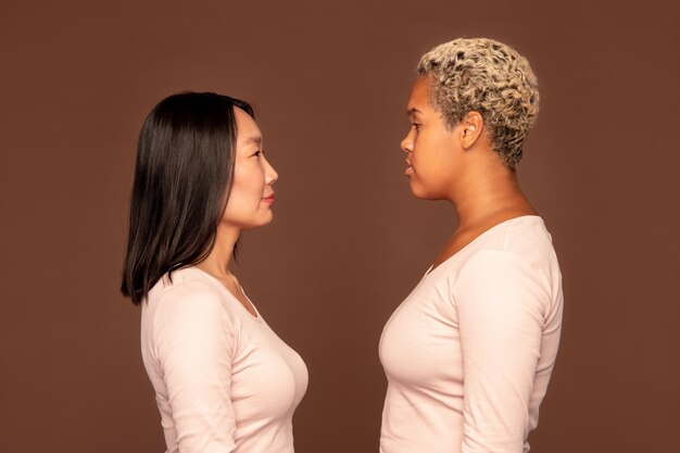 Side view of two young intercultural women in white pullovers standing in front of camera against brown background and looking at one another