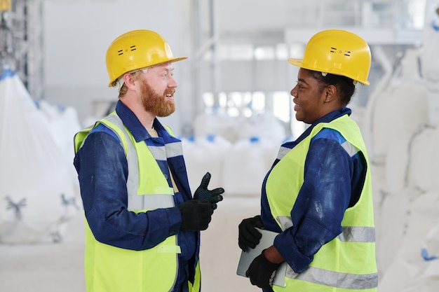 Side view of two young intercultural workers of factory in uniform and hardhats