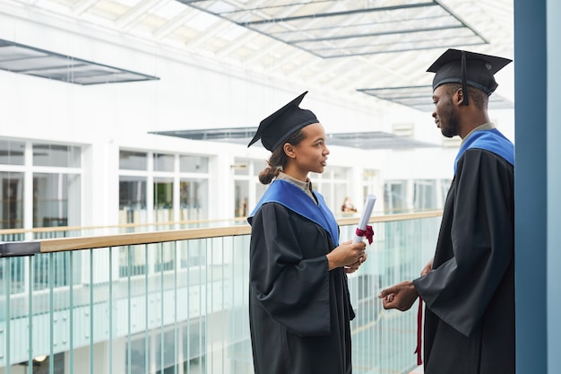Side view at two young people wearing graduation gowns chatting indoors in modern college interior, copy space