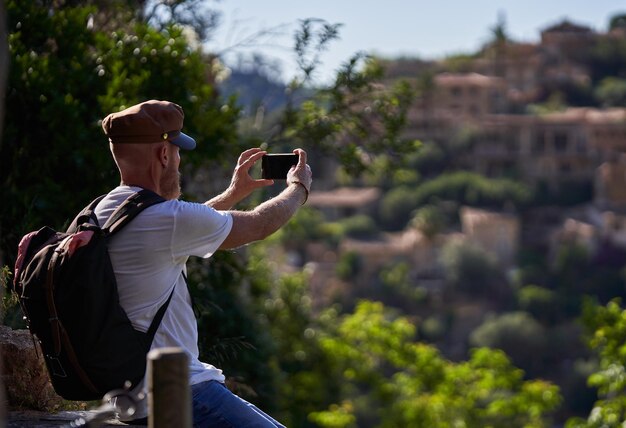 Foto vista laterale di un uomo barbuto irriconoscibile seduto sul recinto in montagne circondate da alberi verdi e scattando foto della città vecchia sul cellulare in una giornata estiva