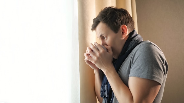 Side view of unwell male in scarf holding cup of aromatic hot beverage while standing near window at home, copy space