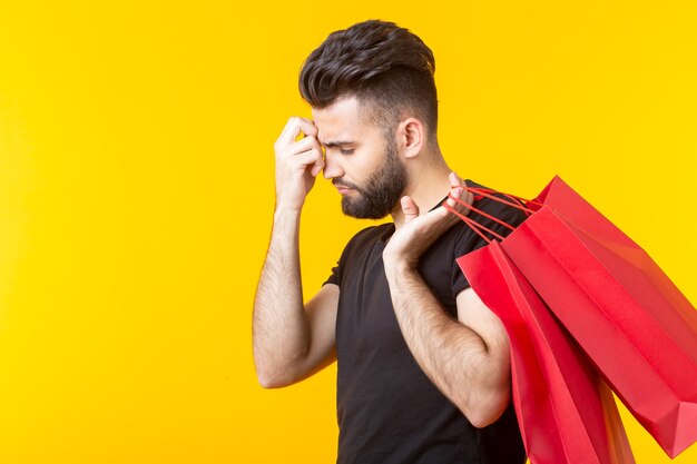 Side view of an upset tired young bearded stylish hipster man holding shopping bags posing on a