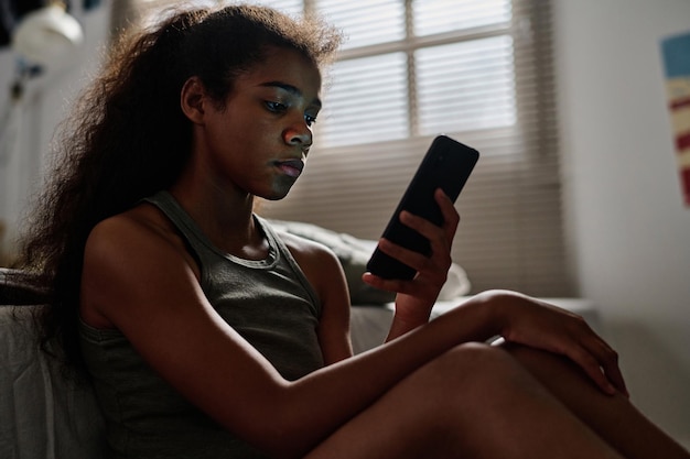 Side view van een wanhopige tiener met een mobiele telefoon die bij het bed in haar kamer zit