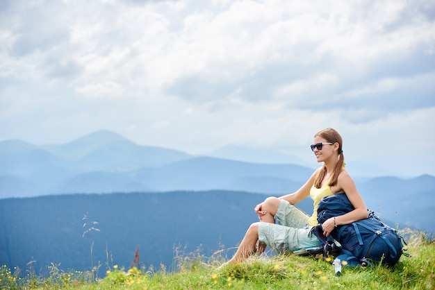 Side view of woman backpacker sitting on grassy hill with backpack, enjoying summer cloudy day in the Carpathian mountains