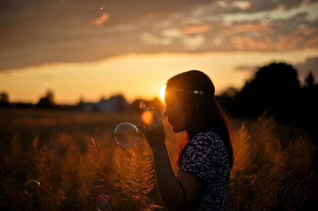 Foto vista laterale di una donna che soffia bolle mentre è in piedi sul campo durante il tramonto