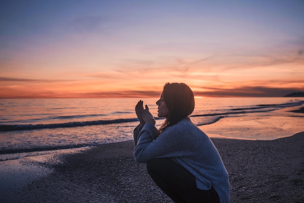 Photo side view of woman crouching at beach against sky during sunset