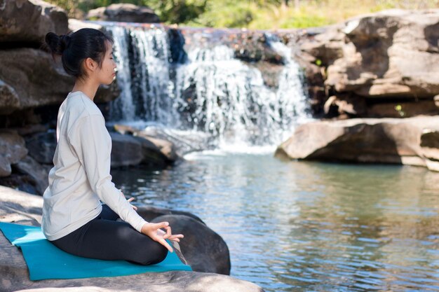 Side view of woman doing lotus position yoga while sitting on rocks against waterfall