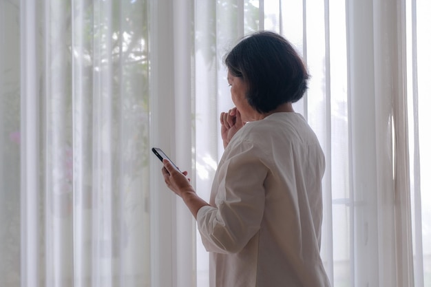 Side view A woman holding a cell phone standing near the window biting her nails