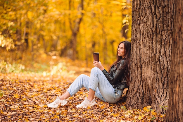 Side view of woman holding coffee cup sitting by tree trunk