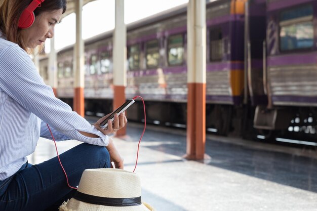 Photo side view of woman listening music over mobile phone while sitting on bench at railroad station platform