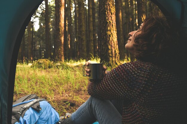 Photo side view of woman looking up while holding coffee mug in tent at forest