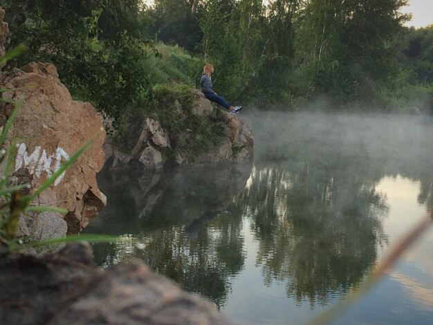 Side view of woman sitting on rock by lake during foggy weather