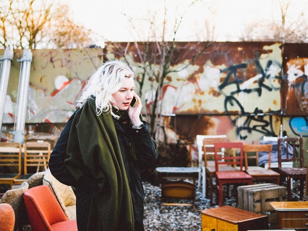 Photo side view of woman talking on smart phone surrounded by chairs in flea market