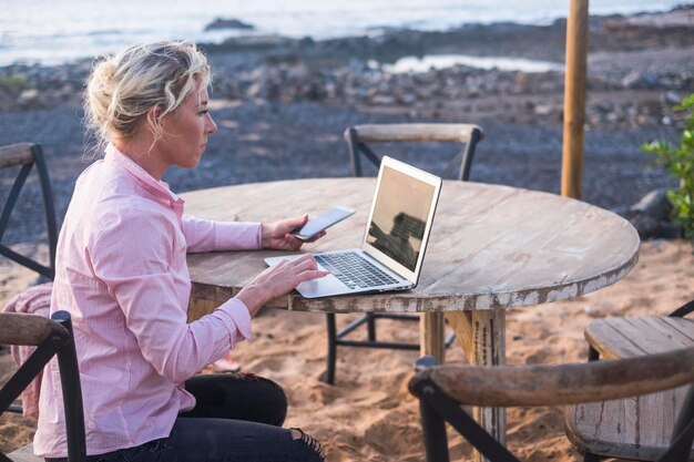 Photo side view of woman using laptop and mobile phone while sitting on table at beach