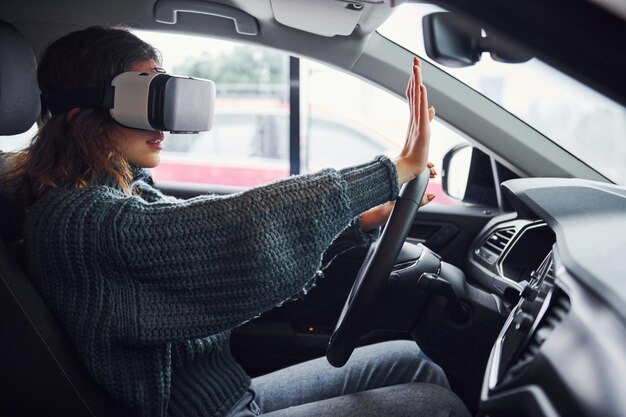 Photo side view of woman in virtual reality glasses that sitting inside of modern car.