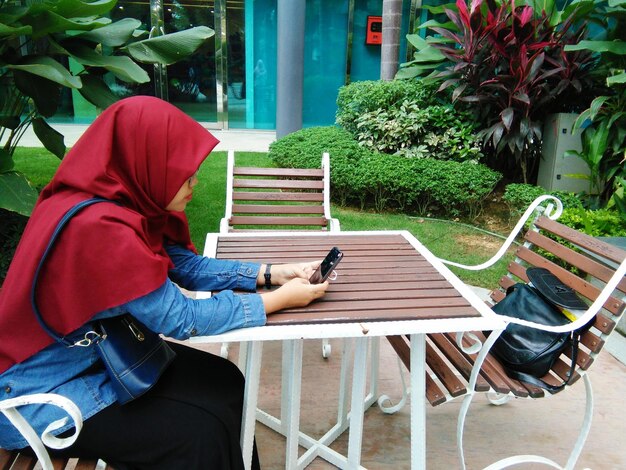 Side view of woman wearing hijab using smart phone while sitting outdoors