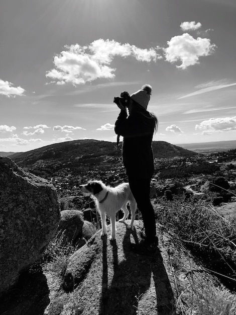 Foto vista laterale di una donna con un cane che fotografa mentre è in piedi sulla montagna contro il cielo